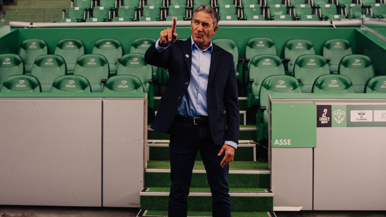 Philippe Montanier, en costume, photographié devant les sièges du banc stéphanois, au bord de la pelouse du Stade Geoffroy-Guichard.
