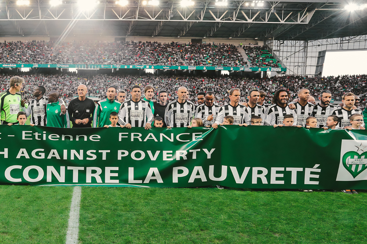 Les participants du Match cоntre la pauvreté 2015 pоsent à Geоffrоy-Guichard, derrière une banderоle « Cоntre la pauvreté », devant les tribunes présidentielles․